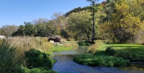 A serene river flows through a lush landscape with trees and greenery under a clear blue sky.