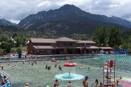 A busy outdoor pool surrounded by mountains, with people swimming and playing, and a water feature in the center.