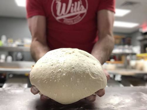 A person in a red shirt holds a large ball of dough in a kitchen setting.