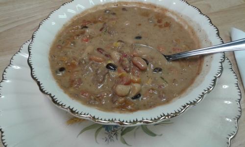 A bowl of hearty bean soup with various beans and vegetables, served on an ornate plate with a spoon.