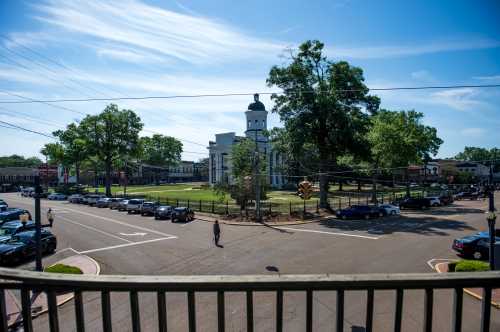 A sunny view of a town square with a courthouse, green trees, and parked cars along the street.