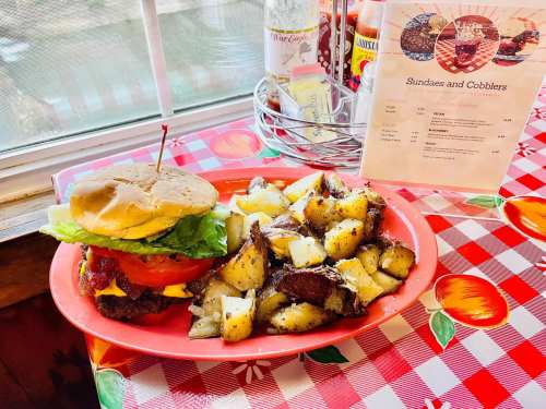 A plate with a burger topped with lettuce and tomato, served with seasoned potato wedges, on a checkered tablecloth.