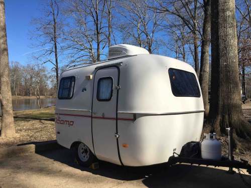 A small white camper trailer parked near a lake, surrounded by bare trees and a clear blue sky.