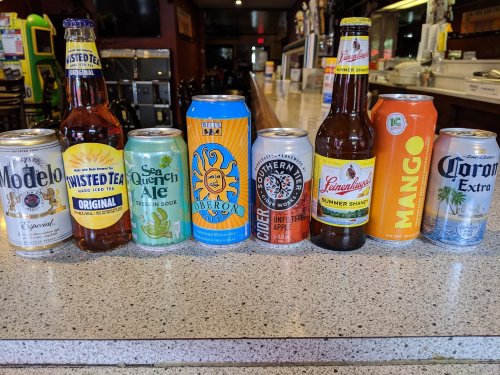 A variety of beer cans and bottles lined up on a bar counter, including Modelo, Twisted Tea, and Corona Extra.