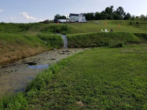 A small house on a hill overlooking a grassy area and a stream with chairs nearby under a clear blue sky.
