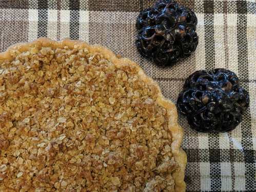 A golden-brown crumble pie on a checkered cloth, accompanied by two dark decorative pumpkins.