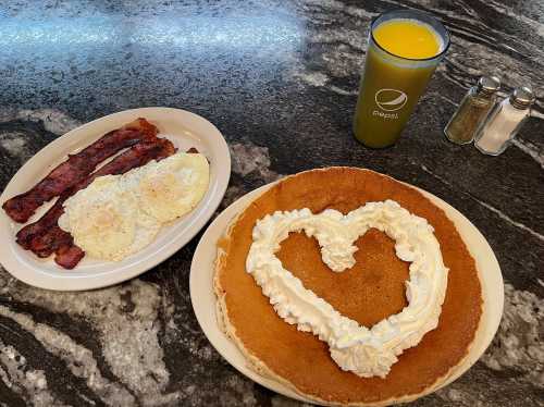 A plate with heart-shaped whipped cream pancake, eggs, bacon, and a glass of orange juice on a marble table.