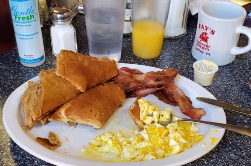 A plate with scrambled eggs, bacon, and broken pancakes, alongside a glass of orange juice and a coffee mug.