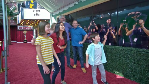 A group of four people, including two children, pose happily in front of a red carpet and photographers.