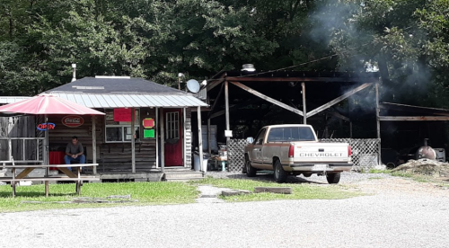 A rustic roadside stand with a man sitting outside, a Chevrolet truck parked nearby, and smoke rising from a grill.