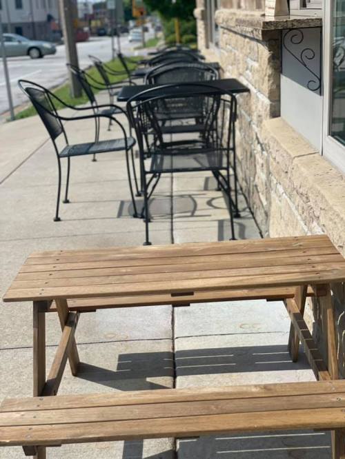 A wooden table on a sidewalk next to empty black chairs outside a building on a sunny day.