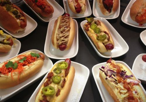A variety of gourmet hot dogs on white plates, topped with colorful ingredients like jalapeños, carrots, and slaw.