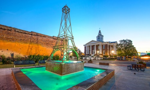 A green-lit fountain resembling the Eiffel Tower, with a historic building and trees in the background at dusk.