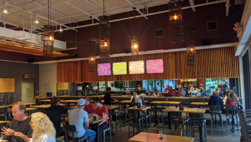 A modern restaurant interior with wooden accents, hanging lights, and patrons dining at tables. Screens display menu items.