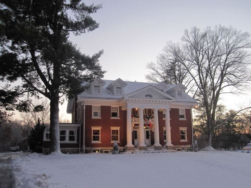 A large brick mansion with white columns, surrounded by snow and trees, under a clear sky at dusk.