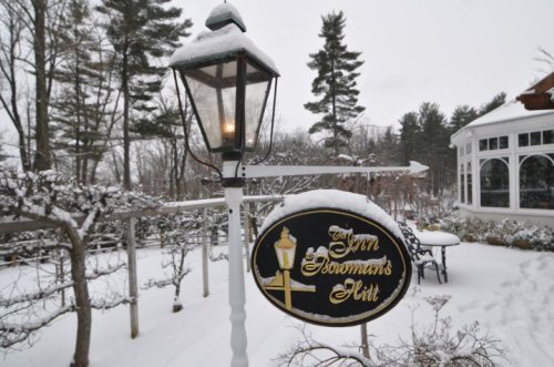 A snowy scene featuring a lamp post and a sign for "Gourman's Hill" surrounded by trees and a winter landscape.