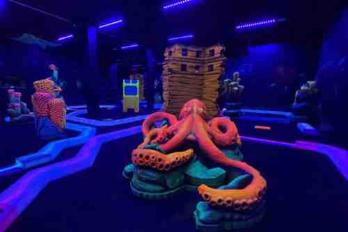 A vibrant, neon-lit indoor space featuring an octopus sculpture and various colorful structures against a dark background.