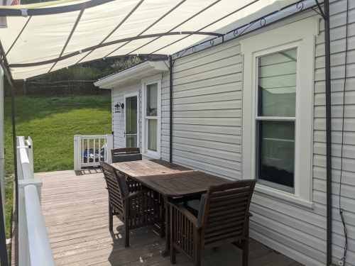 A wooden patio with a table and chairs, shaded by a canopy, next to a house and green lawn.