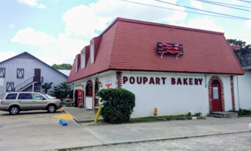 A bakery with a red roof and white walls, featuring a sign that reads "Poupart Bakery," surrounded by a parking area.