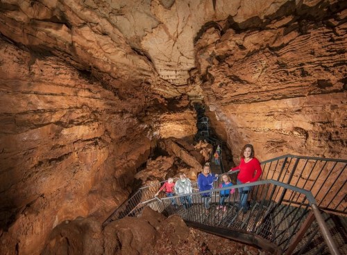 A group of people stands on a metal staircase inside a cave, surrounded by rocky walls and natural formations.
