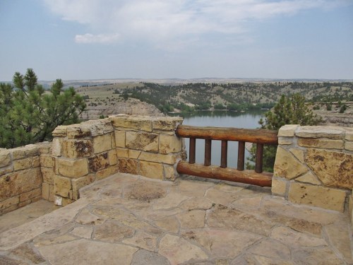 Scenic overlook with stone walls and wooden railing, featuring a view of a lake and surrounding hills.