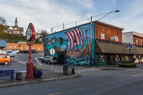 Colorful mural of a ship on a building, with a vintage clock and parked cars in a quaint town setting.