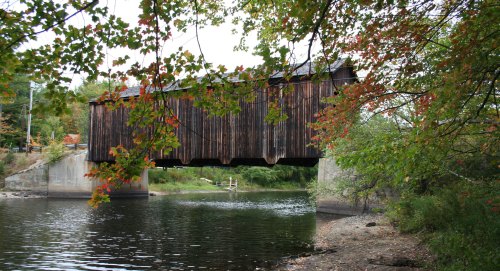 A wooden covered bridge spans a calm river, framed by colorful autumn leaves.