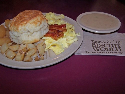 A plate of breakfast featuring a biscuit, scrambled eggs, bacon, potatoes, and a side of gravy, with a napkin from Tudor's Biscuit World.