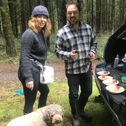 A woman and a man stand by a vehicle in a forest, with a dog nearby and food laid out on a table.