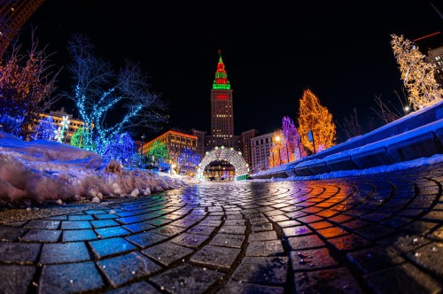 A vibrant cityscape at night, featuring colorful holiday lights and a snowy pathway leading to a tall, illuminated tower.
