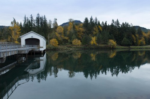 A serene scene of a white covered bridge reflecting in calm water, surrounded by autumn trees and mountains.