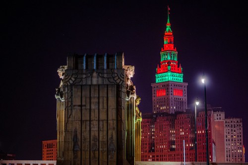 A nighttime cityscape featuring a brightly lit tower in red and green, with a decorative building in the foreground.