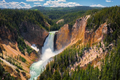 A stunning waterfall cascades into a canyon, surrounded by lush green forests and rocky cliffs under a blue sky.