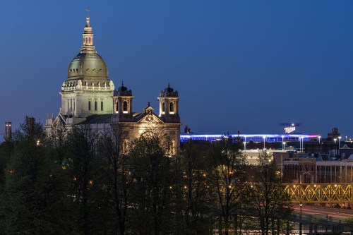 A historic building with a large dome and towers, illuminated at dusk, surrounded by trees and modern structures.
