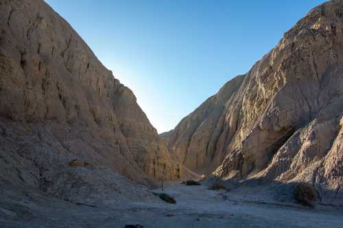 A narrow canyon with steep, rocky walls under a clear blue sky, showcasing rugged terrain and natural formations.