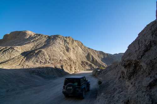 A black Jeep drives along a dusty road surrounded by rugged, rocky hills under a clear blue sky.