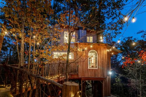 A cozy treehouse illuminated by string lights, surrounded by autumn foliage at dusk.