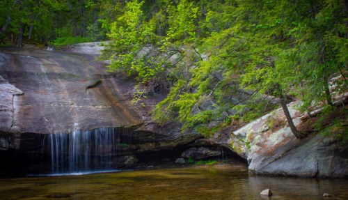 A serene waterfall cascades over rocks into a calm pool, surrounded by lush green trees and a rocky landscape.