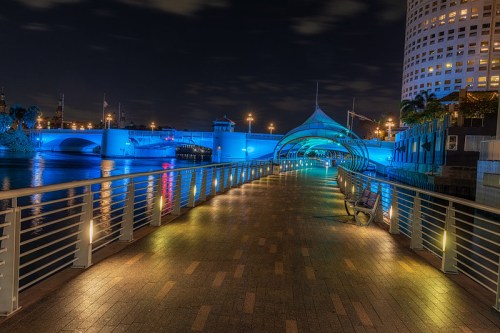 A lit walkway along a river at night, featuring blue lights and a modern archway structure.