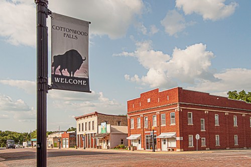 A welcome banner for Cottonwood Falls stands beside historic brick buildings under a blue sky with fluffy clouds.