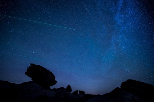 A starry night sky filled with stars and streaks of light, silhouetted by dark rock formations in the foreground.