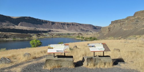 Two informational signs on stone bases overlook a river surrounded by dry grass and rocky cliffs under a clear blue sky.