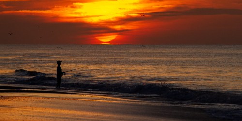 A lone fisherman stands on the shore at sunset, casting a line into the calm ocean waters.