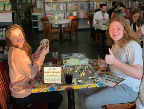 Two friends smile at a table in a game café, holding cards and enjoying drinks, with board games spread out.