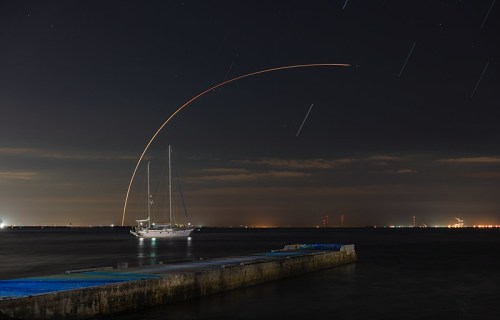 A sailboat anchored near a pier at night, with star trails and city lights in the background.