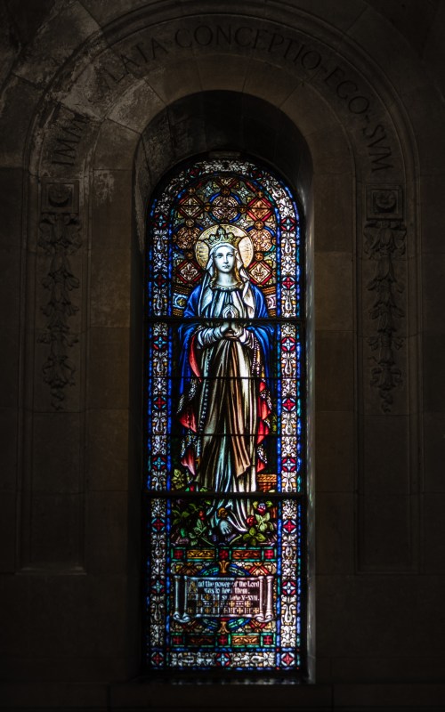 Stained glass window depicting a figure in religious attire, surrounded by colorful patterns and inscriptions.