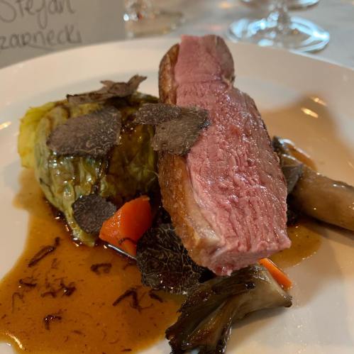 A plated dish featuring sliced pink meat, vegetables, and dark sauce, garnished with herbs and truffle slices.