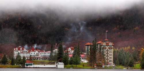 A scenic view of a historic hotel by a lake, surrounded by misty mountains and autumn foliage.