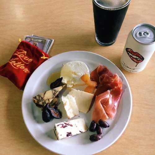 A plate with assorted cheeses, cured meats, dried fruits, olives, and a drink beside a small red pouch.