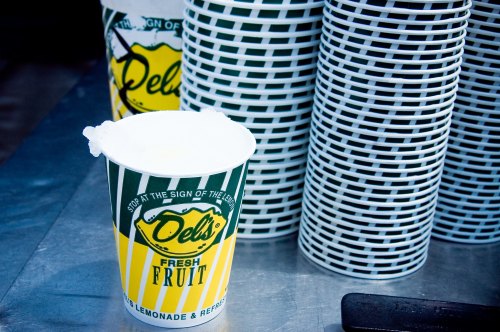 A colorful cup labeled "Dels Fresh Fruit" next to stacks of white cups on a metal surface.